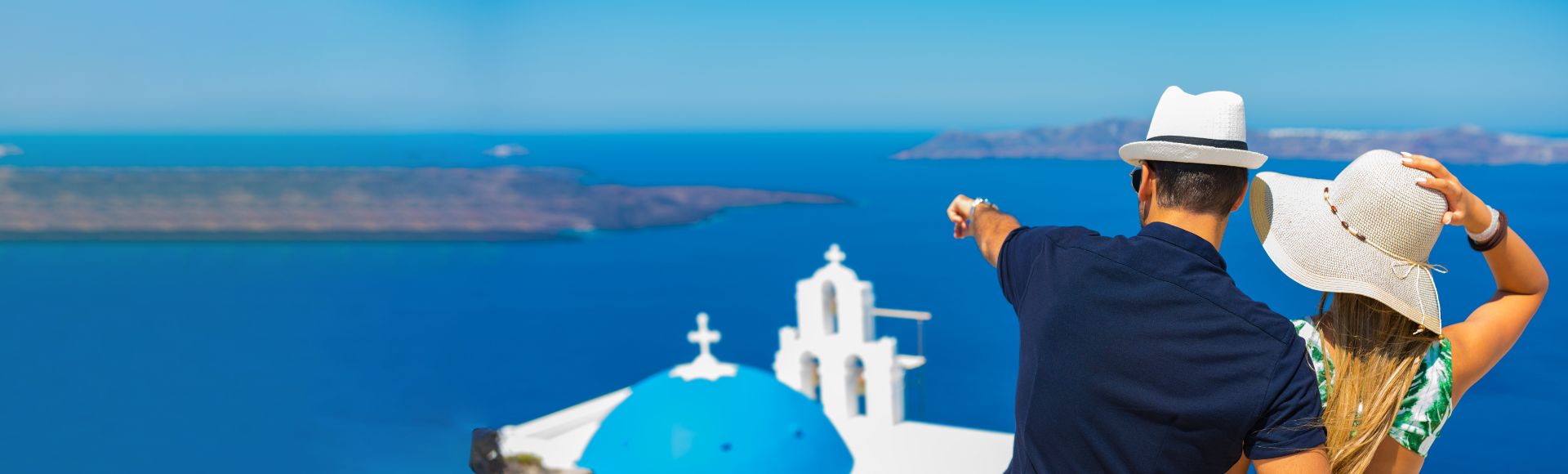 A couple wearing hats sits on a wall overlooking the sea, with white buildings and a blue-domed church in Santorini, Greece, in the background.