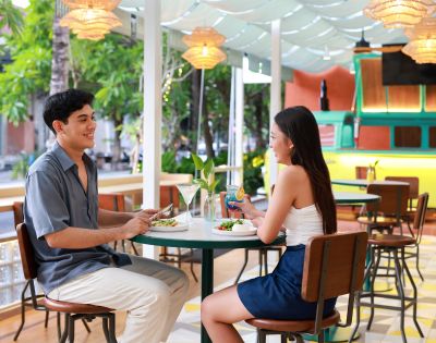 A smiling man and woman sit at an outdoor cafe table, sharing a meal. Surrounded by lush greenery, the setting is relaxed and tropical.