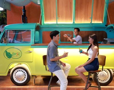 A couple sits on stools enjoying drinks at a stylish bar made from a yellow-green van. A bartender stands inside, smiling, creating a lively atmosphere.