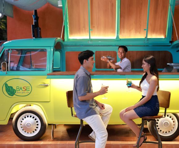 A couple sits on stools enjoying drinks at a stylish bar made from a yellow-green van. A bartender stands inside, smiling, creating a lively atmosphere.