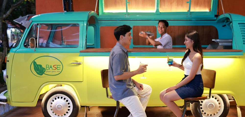 A couple sits on stools enjoying drinks at a stylish bar made from a yellow-green van. A bartender stands inside, smiling, creating a lively atmosphere.