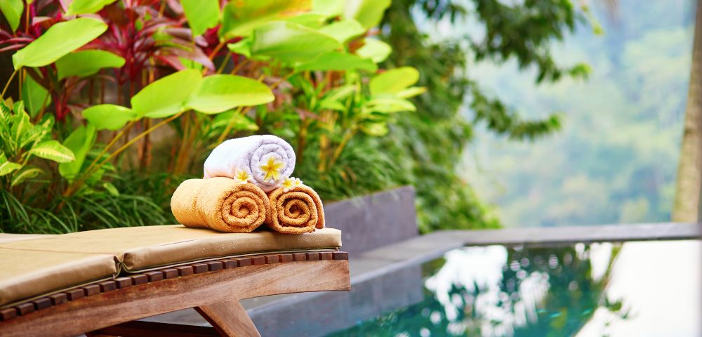 A luxurious poolside scene featuring a wooden lounge chair with two rolled brown towels and a white towel topped with a flower, surrounded by lush greenery.