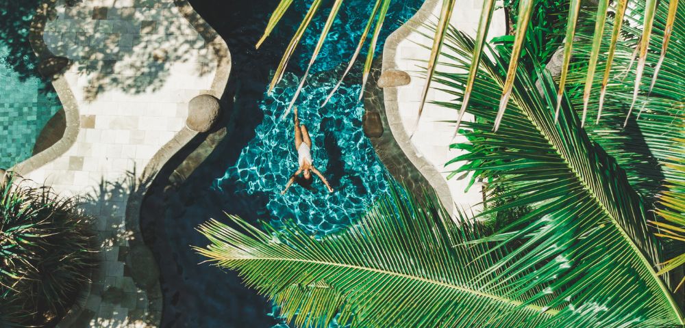 A person floats in a curvy blue pool surrounded by lush green palm leaves and patterned stone pathways, creating a tranquil, tropical vibe.