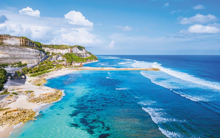 Aerial view of a stunning beach with turquoise water, gentle waves, and rocky cliffs under a clear blue sky, conveying serenity and natural beauty.