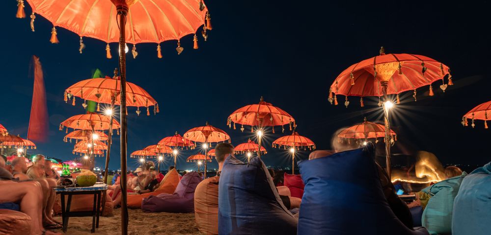 Beach scene at night with vibrant orange umbrellas softly lit, people relaxing on colorful bean bags, creating a warm, inviting atmosphere.