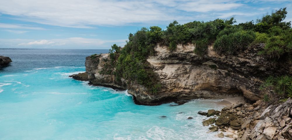 Rocky cliff topped with lush greenery overlooking clear turquoise water on a sunny day. Waves gently crash against the shore, creating a serene scene.