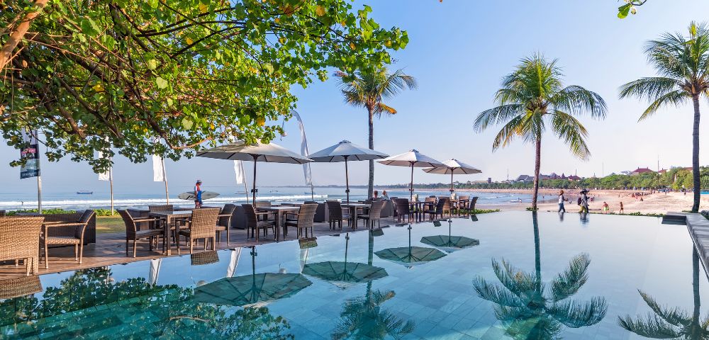 Poolside dining area with wicker chairs and umbrellas overlooks a serene beach with palm trees. Clear blue sky and tranquil reflections create a relaxing vibe.