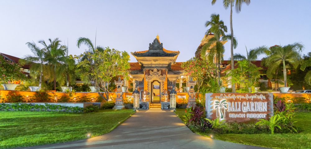 Pathway leading to a well-lit, ornate Balinese entrance at Bali Garden Beach Resort. Surrounded by lush greenery and palm trees at dusk.
