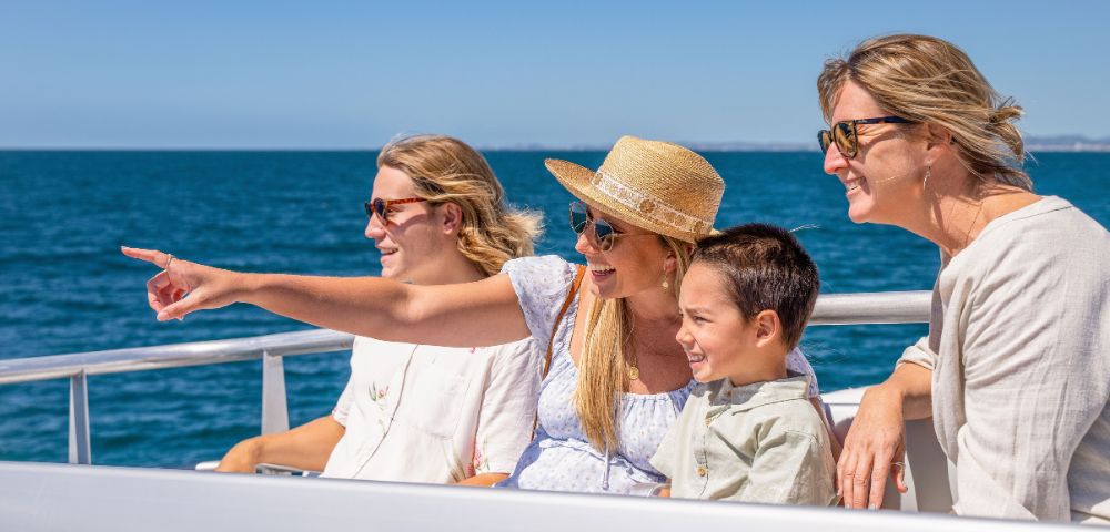 Four people smile and enjoy a sunny day on a boat. One points toward the ocean, highlighting excitement. Blue sea and sky form a serene backdrop.