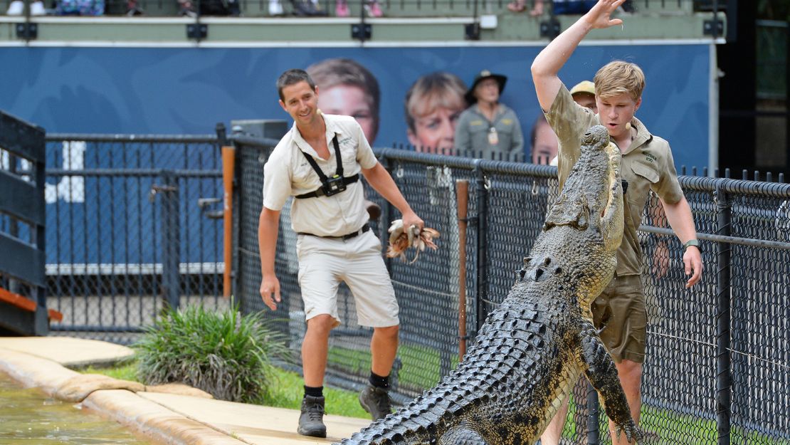 Young man in a khaki uniform feeds a large crocodile beside a fenced area, while another man watches, smiling. Onlookers are visible in the background.