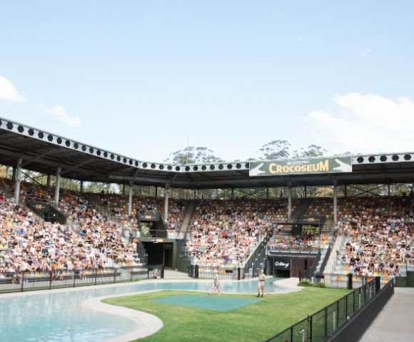 A large crowd seated in an open-air arena, the Crocoseum. The central area features a water pool and grass. The atmosphere is lively and engaging.