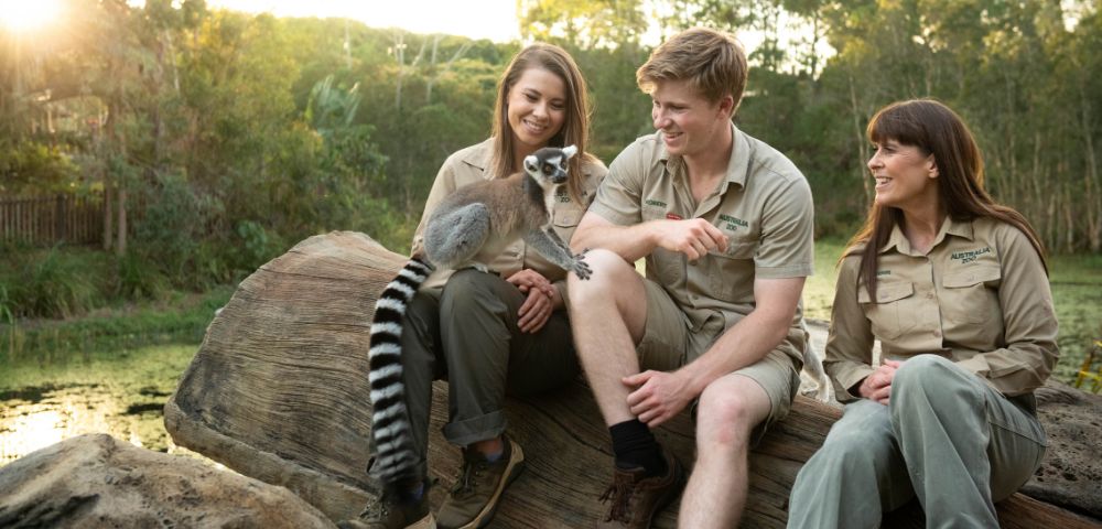 Three people in khaki gear sit on a large log interacting with a lemur. They are outdoors with a pond and trees in the background, smiling warmly.