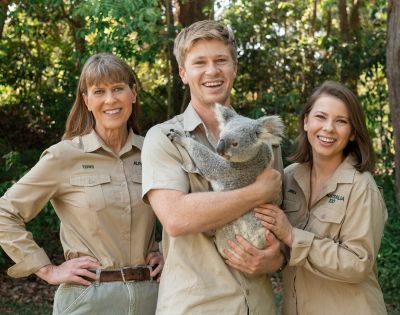 Three people in khaki uniforms smiling outdoors, surrounded by lush greenery. The person in the middle is holding a calm koala, creating a joyful and engaging atmosphere.