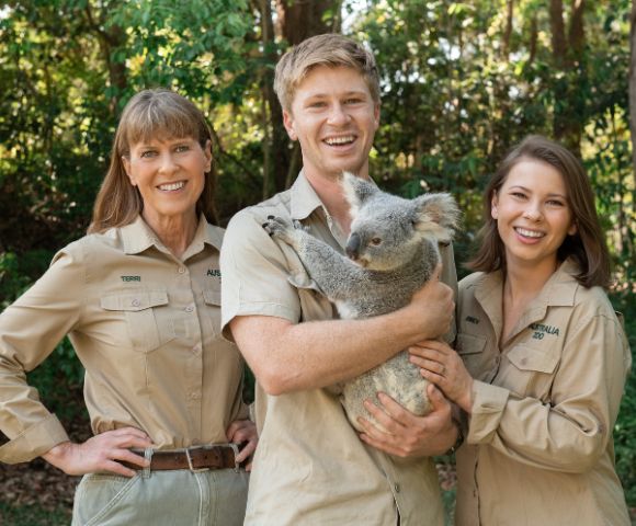 Three people in khaki uniforms smiling outdoors, surrounded by lush greenery. The person in the middle is holding a calm koala, creating a joyful and engaging atmosphere.
