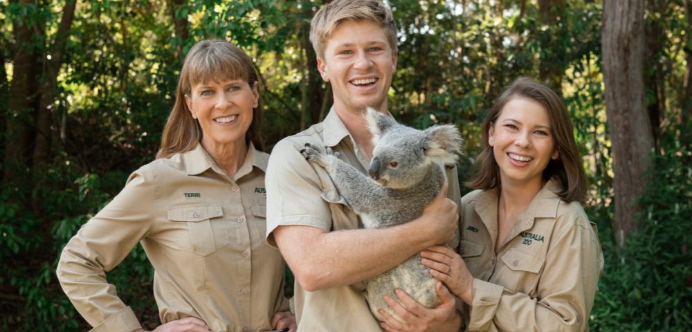 Three people in khaki uniforms smiling outdoors, surrounded by lush greenery. The person in the middle is holding a calm koala, creating a joyful and engaging atmosphere.