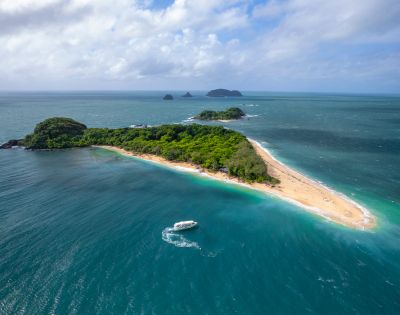 Aerial view of a lush, green island with a long sandy beach surrounded by turquoise ocean. A white boat leaves a wake, suggesting a tranquil setting.