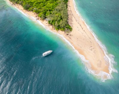 Aerial view of a lush, green island with a narrow sandy beach surrounded by vibrant turquoise waters. A small boat floats near the shore, evoking serenity.