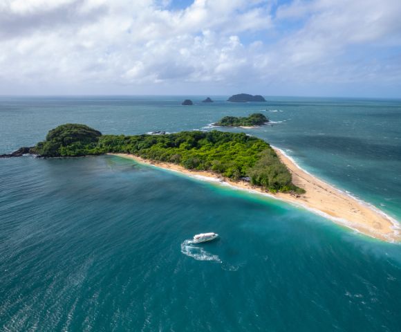 Aerial view of a lush, green island with a long sandy beach surrounded by turquoise ocean. A white boat leaves a wake, suggesting a tranquil setting.