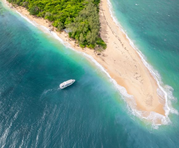 Aerial view of a lush, green island with a narrow sandy beach surrounded by vibrant turquoise waters. A small boat floats near the shore, evoking serenity.