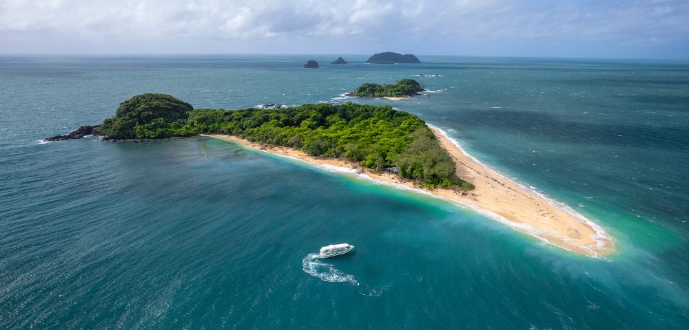 Aerial view of a lush, green island with a long sandy beach surrounded by turquoise ocean. A white boat leaves a wake, suggesting a tranquil setting.