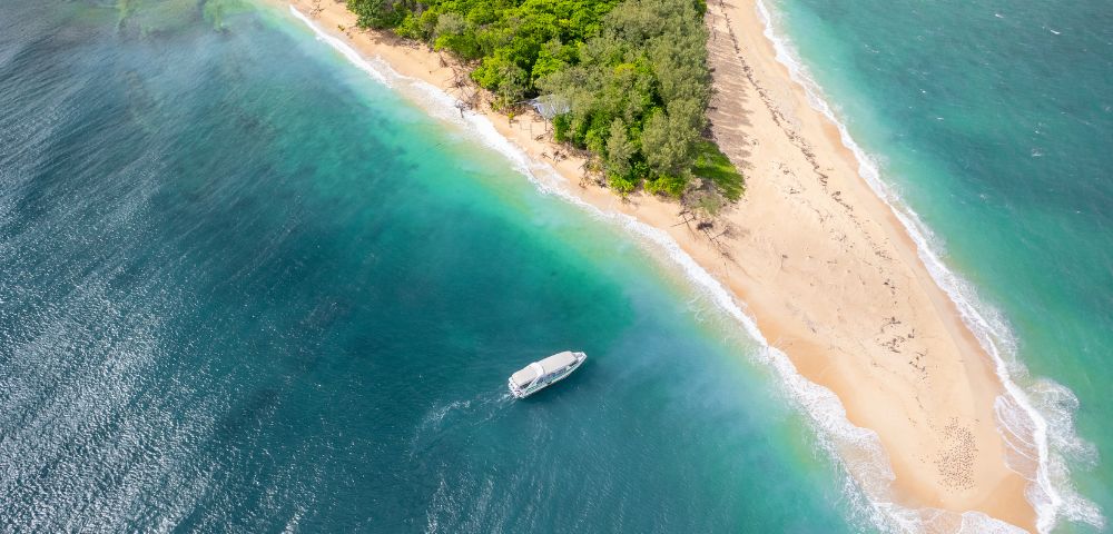 Aerial view of a lush, green island with a narrow sandy beach surrounded by vibrant turquoise waters. A small boat floats near the shore, evoking serenity.