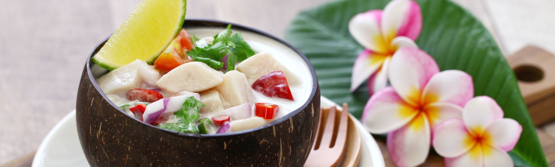 A coconut bowl of ceviche with lime garnishes, set on a white plate. Wooden utensils and pink plumeria flowers on a large green leaf create a fresh, tropical feel.