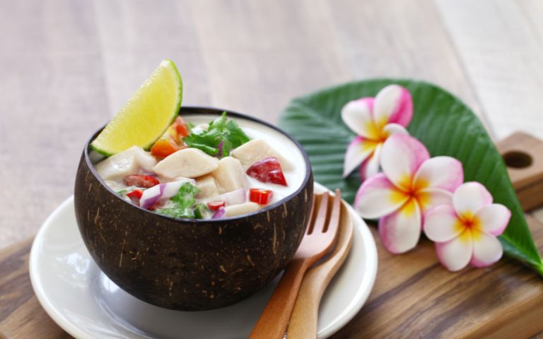 A coconut bowl of ceviche with lime garnishes, set on a white plate. Wooden utensils and pink plumeria flowers on a large green leaf create a fresh, tropical feel.