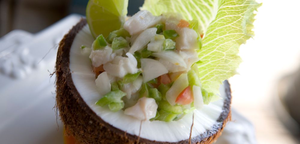 Ceviche served in a halved coconut shell, garnished with lime and lettuce, featuring diced fish, onions, and peppers; fresh and tropical presentation.