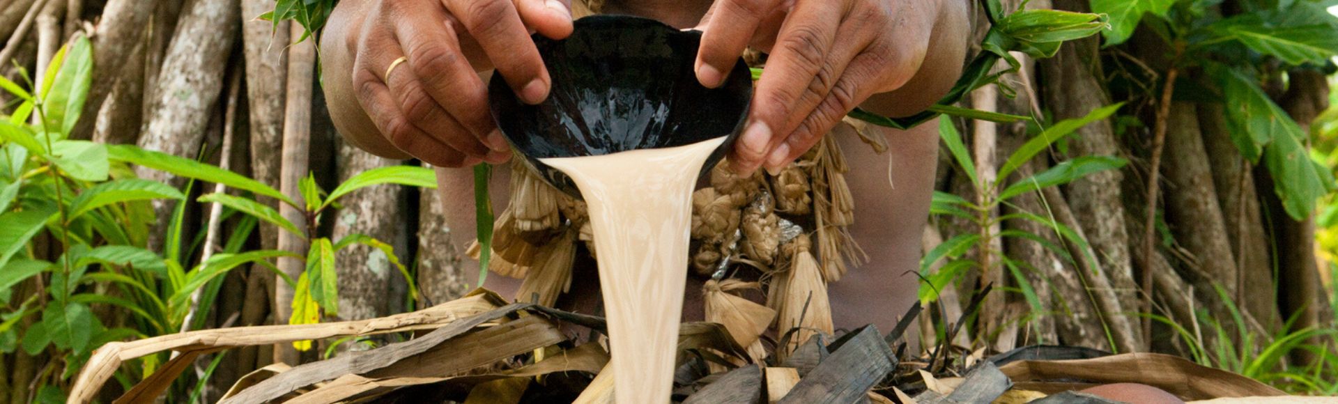 A man wearing traditional attire pours liquid from a black bowl. He is surrounded by lush greenery and large tree roots, conveying a cultural ritual.