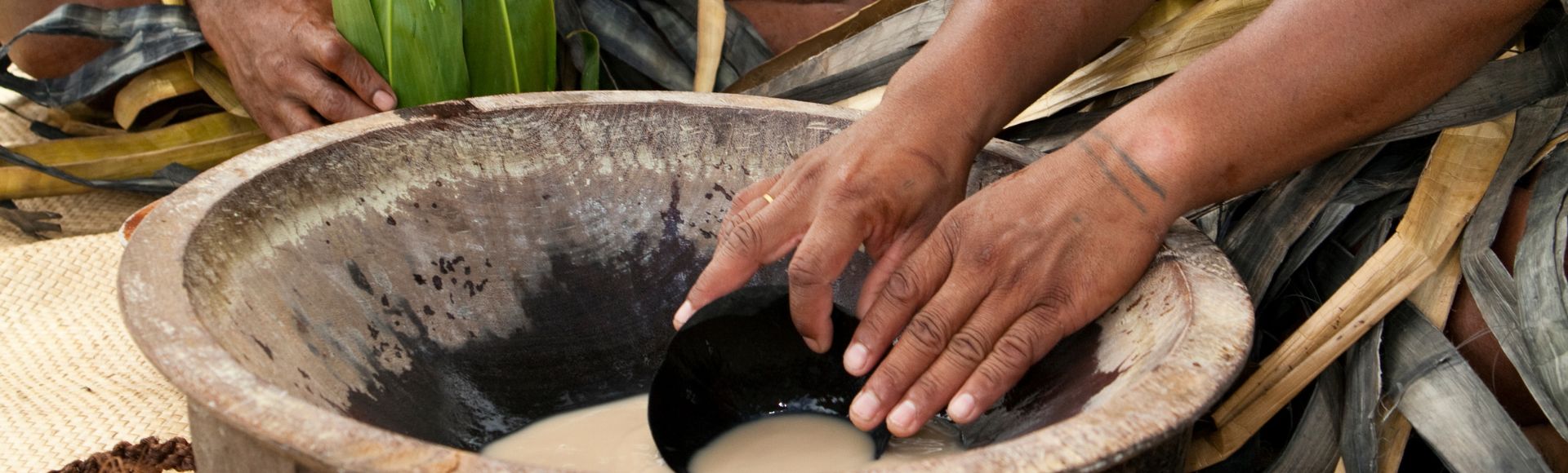 Hands preparing traditional kava in a wooden bowl, surrounded by woven mats and leaves, conveying cultural richness and ceremonial practice.