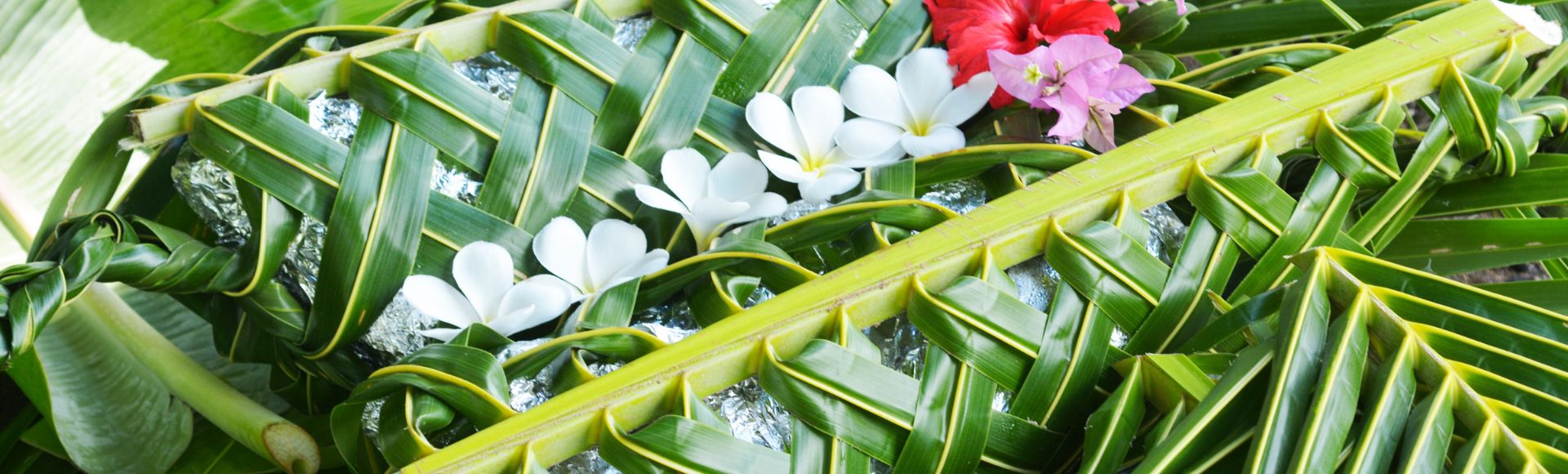 A woven green palm leaf basket adorned with white and red tropical flowers, resting on large green leaves. The scene conveys a natural, tropical vibe.