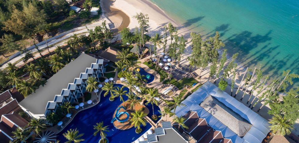 Aerial view of a tropical beach resort with turquoise sea, sandy beach, palm trees, and a curvy pool surrounded by sun loungers and villas. Peaceful ambiance.