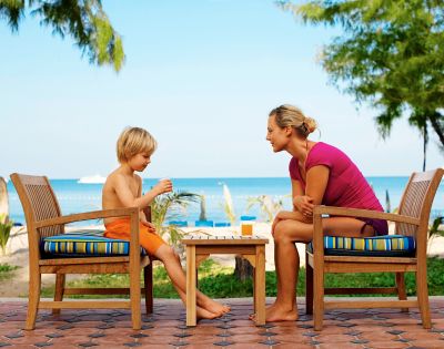 A child and an adult sit outdoors on wooden chairs by a beach, enjoying drinks. The scene is bright and relaxed, suggesting a warm, sunny day.