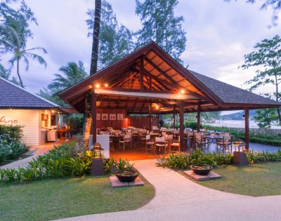 Open-air restaurant with wooden roof, surrounded by lush greenery. Tables and chairs are set under warm lighting, creating a cozy, inviting atmosphere.