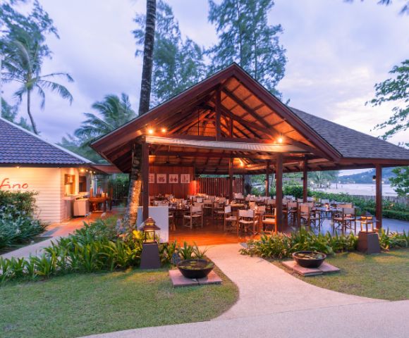 Open-air restaurant with wooden roof, surrounded by lush greenery. Tables and chairs are set under warm lighting, creating a cozy, inviting atmosphere.