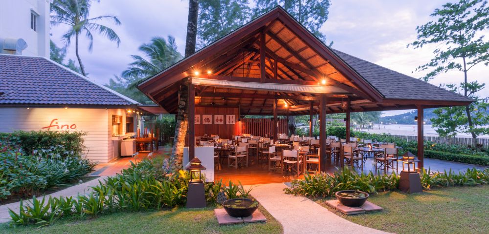 Open-air restaurant with wooden roof, surrounded by lush greenery. Tables and chairs are set under warm lighting, creating a cozy, inviting atmosphere.