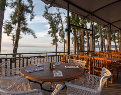Outdoor restaurant with a wooden table set for dining, surrounded by wicker chairs. Overlooks a serene beach and tranquil ocean under a dimming sun.