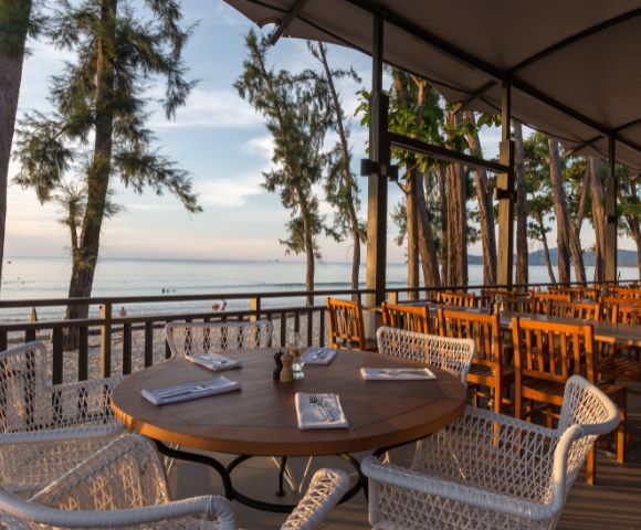 Outdoor restaurant with a wooden table set for dining, surrounded by wicker chairs. Overlooks a serene beach and tranquil ocean under a dimming sun.