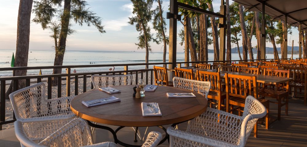Outdoor restaurant with a wooden table set for dining, surrounded by wicker chairs. Overlooks a serene beach and tranquil ocean under a dimming sun.