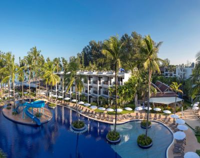 Tropical resort with large lagoon-style pool, surrounded by palm trees and sun loungers. Modern white buildings and clear blue sky create a serene atmosphere.
