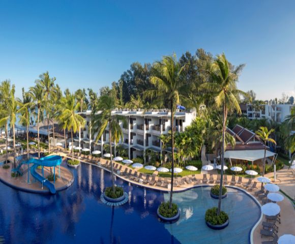 Tropical resort with large lagoon-style pool, surrounded by palm trees and sun loungers. Modern white buildings and clear blue sky create a serene atmosphere.