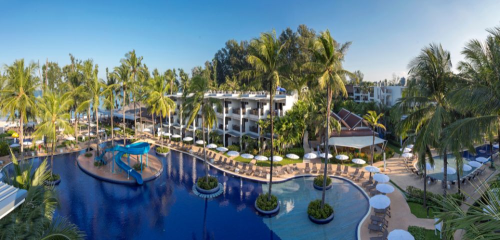Tropical resort with large lagoon-style pool, surrounded by palm trees and sun loungers. Modern white buildings and clear blue sky create a serene atmosphere.