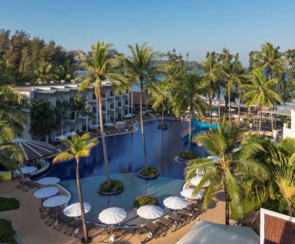 Aerial view of a tropical resort featuring a large, curvy swimming pool surrounded by palm trees, lounge chairs, and umbrellas, with beach and sea in the background.