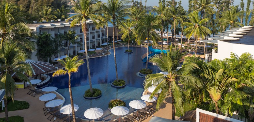 Aerial view of a tropical resort featuring a large, curvy swimming pool surrounded by palm trees, lounge chairs, and umbrellas, with beach and sea in the background.