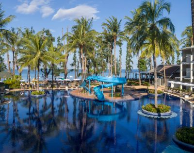 Tropical resort pool with palm trees, blue waterslide, and lounge chairs under umbrellas. Ocean view in the background under a clear blue sky.