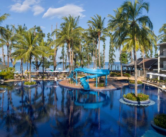 Tropical resort pool with palm trees, blue waterslide, and lounge chairs under umbrellas. Ocean view in the background under a clear blue sky.