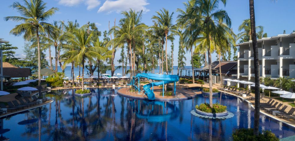Tropical resort pool with palm trees, blue waterslide, and lounge chairs under umbrellas. Ocean view in the background under a clear blue sky.