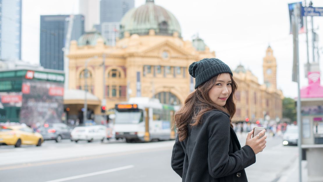 Woman in a black coat and beanie stands on a busy city street, holding a phone. Iconic building and tram in the background. Daytime, lively urban vibe.