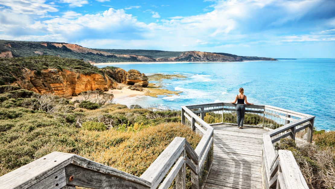 A woman stands on a wooden boardwalk overlooking a stunning coastal view. The scene features cliffs, lush green shrubs, and shimmering blue ocean under a clear sky.