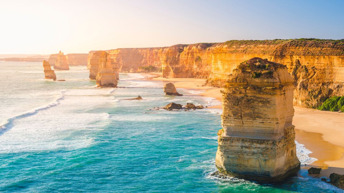 Sunlit coastal scene of the Twelve Apostles in Australia. Eroded limestone stacks rise from blue ocean waves with a backdrop of rugged cliffs.